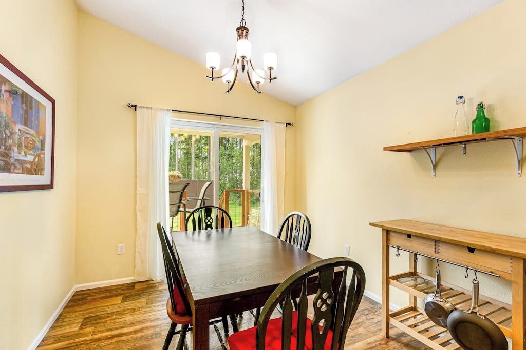 Un coin repas avec une table et des chaises en bois, un lustre, une fenêtre avec des rideaux et une étagère contenant des ustensiles de cuisine.