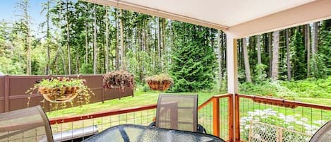 A covered patio with a glass table and chairs, surrounded by a wooden fence and a view of a forest.