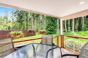 A covered patio with a glass table and chairs, surrounded by a wooden fence and a view of a forest.