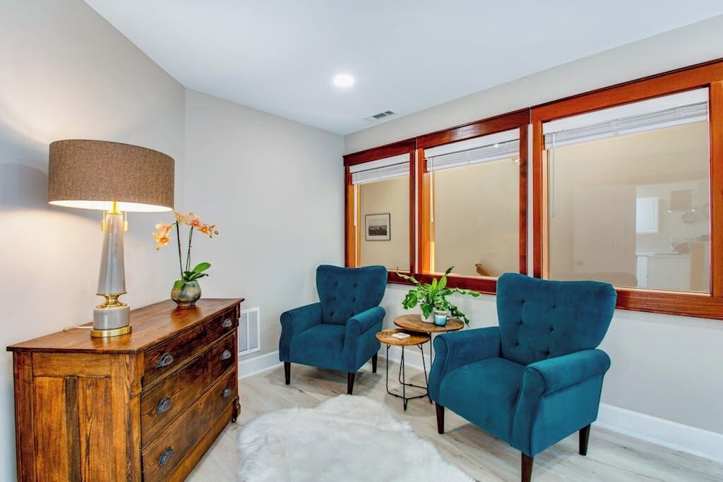 A room with a wooden cabinet, two blue armchairs, a small round table, a lamp, and a mirror with a wooden frame.