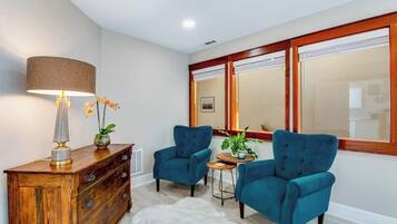 A room with a wooden cabinet, two blue armchairs, a small round table, a lamp, and a mirror with a wooden frame.