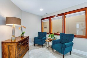 A room with a wooden cabinet, two blue armchairs, a small round table, a lamp, and a mirror with a wooden frame.