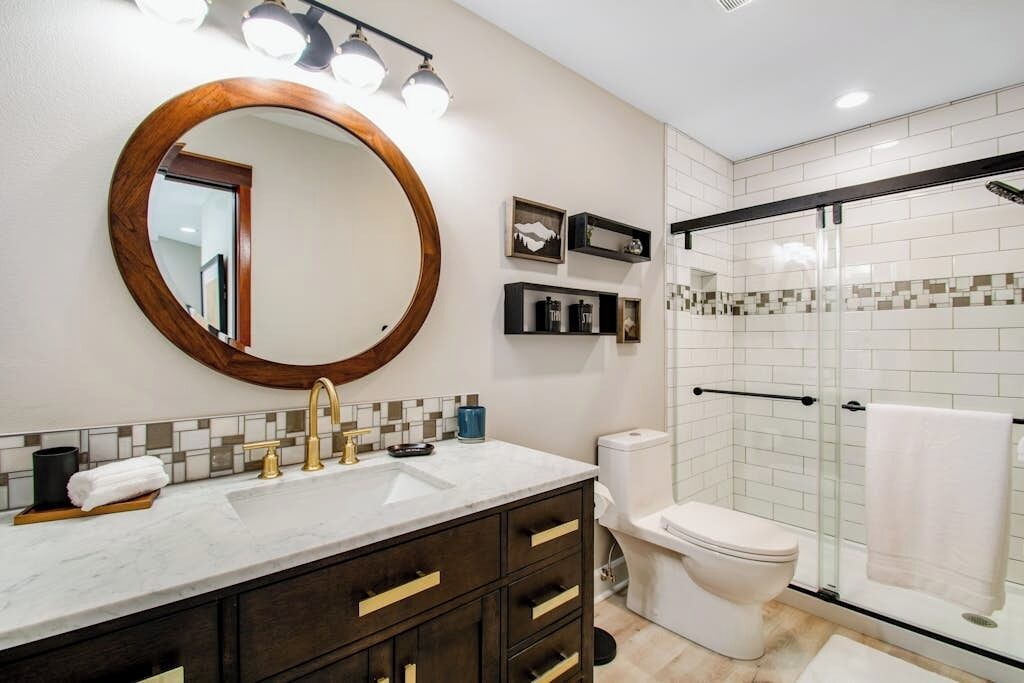 A bathroom with a round mirror, a sink with a gold faucet, a toilet, and a glass shower enclosure.