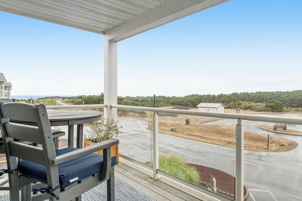 En balkon med udsigt til en vej og grøn bevoksning, et bord med en stol og et gelænder.