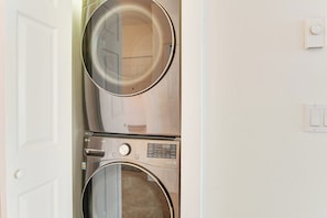 A stacked washer and dryer unit in a room with a white door and light switch on the wall. - Oceanfront 2BR 3rd-Floor | Balcony | Pool (Westport)