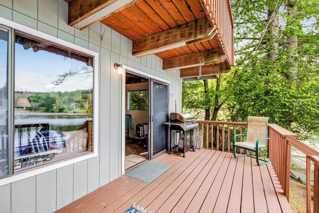 A wooden deck with a grill and a chair, a large window overlooking a body of water, and a door leading to an interior space.