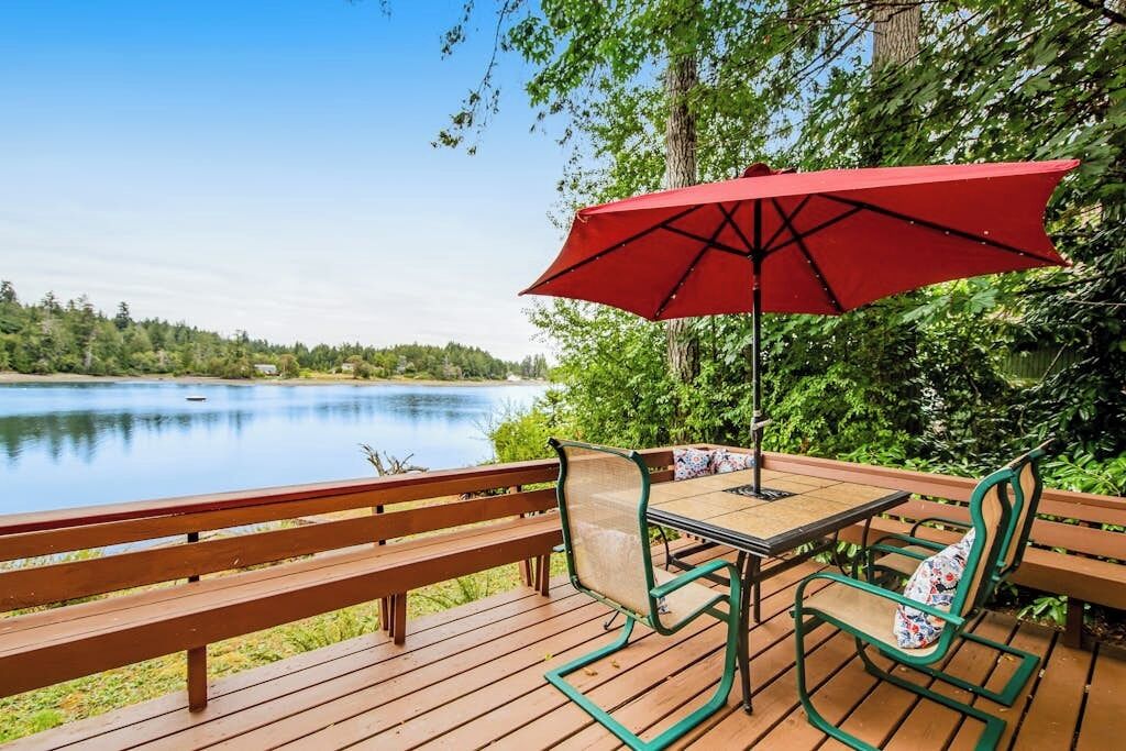 A wooden deck with a table and chairs, a red umbrella, and a view of a lake.
