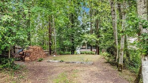 A forested area with a dirt path leading to a building. There is a stack of firewood and a white structure in the background. - 1BR Soundview Dog Friendly - Dock - WoodStove (Shelton)