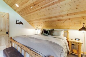 Ein Schlafzimmer mit Holzboden, einem Bett mit grauer Bettdecke, einem Nachttisch mit Lampe und einem an der Wand befestigten Regal mit einem Bild.