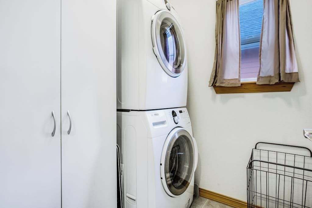 A laundry room with a stacked washer and dryer, a window with curtains, and a metal drying rack.