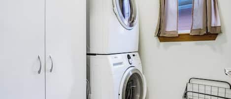 A laundry room with a stacked washer and dryer, a window with curtains, and a metal drying rack.