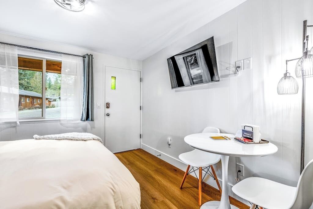 A modern bedroom with a bed, a small round table with chairs, a TV mounted on the wall, and a window with curtains.
