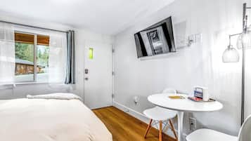 A modern bedroom with a bed, a small round table with chairs, a TV mounted on the wall, and a window with curtains.