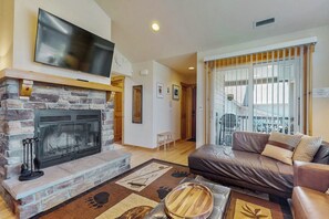 A living room with a stone fireplace, a leather sofa, and a large TV mounted above the fireplace. - 2BR mountainview condo with balcony & pool (Ronald)
