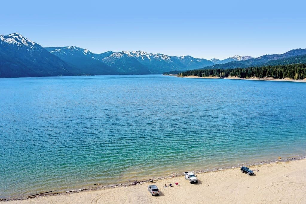 A lake with mountains in the background and a sandy beach with parked vehicles.