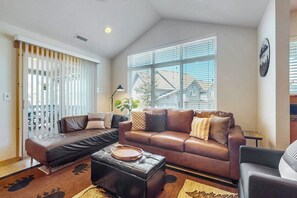 A living room with a brown leather sofa, a black ottoman, and a coffee table. There is a window with blinds, a plant, and a wall-mounted light fixture. - 2BR mountainview condo with balcony & pool (Ronald)