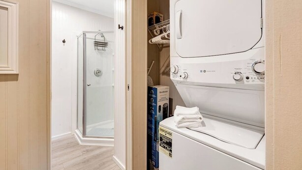 Un cuarto de lavandería con lavadora y secadora de carga frontal, cabina de ducha con mampara de vidrio y un espacio de almacenamiento con ropa colgada.