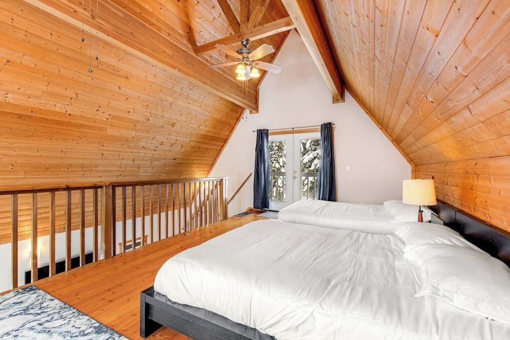 Ein Schlafzimmer mit Holzboden, einem Bett mit weißer Bettwäsche, einem Nachttisch mit Lampe und einem Fenster mit Vorhängen.