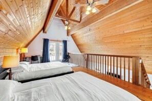 Ein Zimmer mit schrägem Dach, Holzwänden, einem Bett mit weißer Bettwäsche, einem Schreibtisch mit Stuhl und einem Fenster mit Vorhängen.