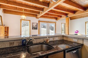 Eine Küche mit Granit-Arbeitsplatten, einer Edelstahl-Spüle, Holzbalken an der Decke und einem Fenster mit Blick auf schneebedeckte Bäume.