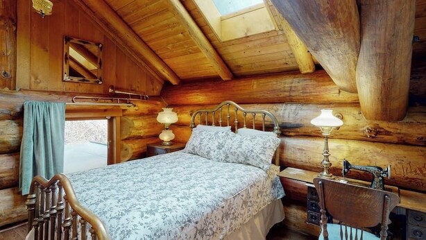 Quarto de cabana de toras com uma cama, uma cadeira, uma máquina de costura e uma janela.