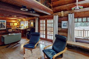 Interior d'una cabana de fusta amb zona de menjador, dues butaques de cuir, taula de fusta i finestra gran amb cortines.