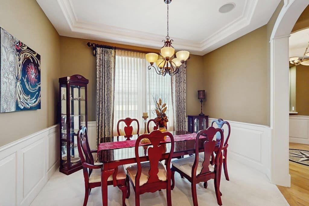 A dining room with a wooden table, chairs, a chandelier, and a large window with curtains.