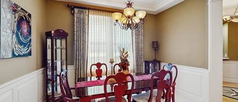 A dining room with a wooden table, chairs, a chandelier, and a large window with curtains.