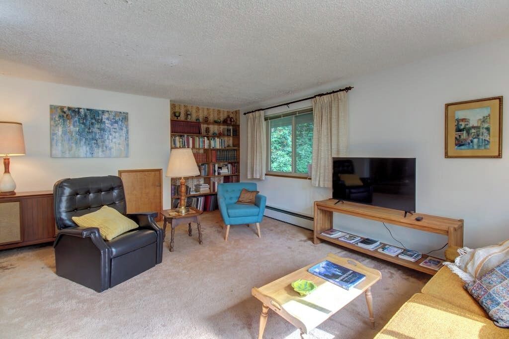 A living room with a black leather sofa, a blue armchair, a wooden coffee table, a bookshelf, a television, and a window with curtains.