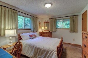 Ein Schlafzimmer mit einem Bett, einem Nachttisch, einer Lampe, einer Kommode und zwei Fenstern mit Vorhängen.