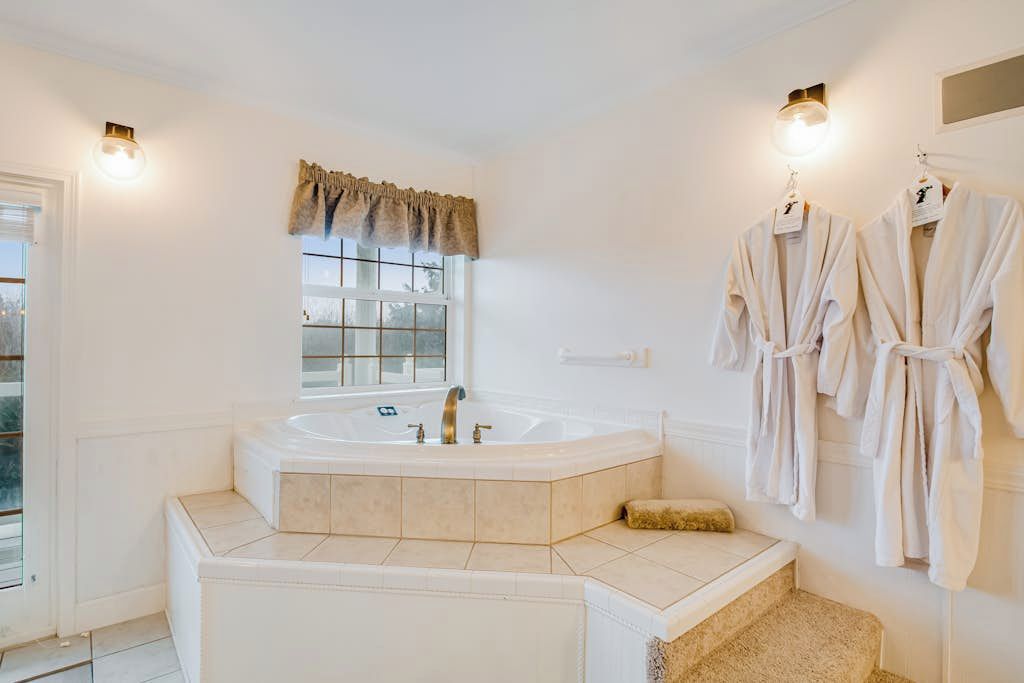 Une salle de bain avec une grande baignoire, une fenêtre avec un rideau et deux peignoirs accrochés au mur.
