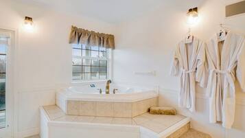 A bathroom with a large bathtub, a window with a curtain, and two robes hanging on the wall.