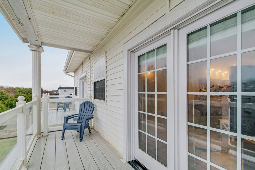 A covered porch with a white railing, a blue chair, and a large window with a view of the interior.