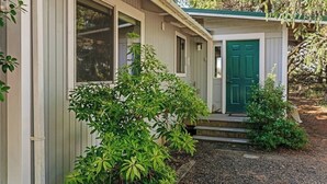 Ein kleines, eingeschossiges Gebäude mit einer grünen Tür, einer Veranda mit Treppen und einem kleinen Garten mit Sträuchern.