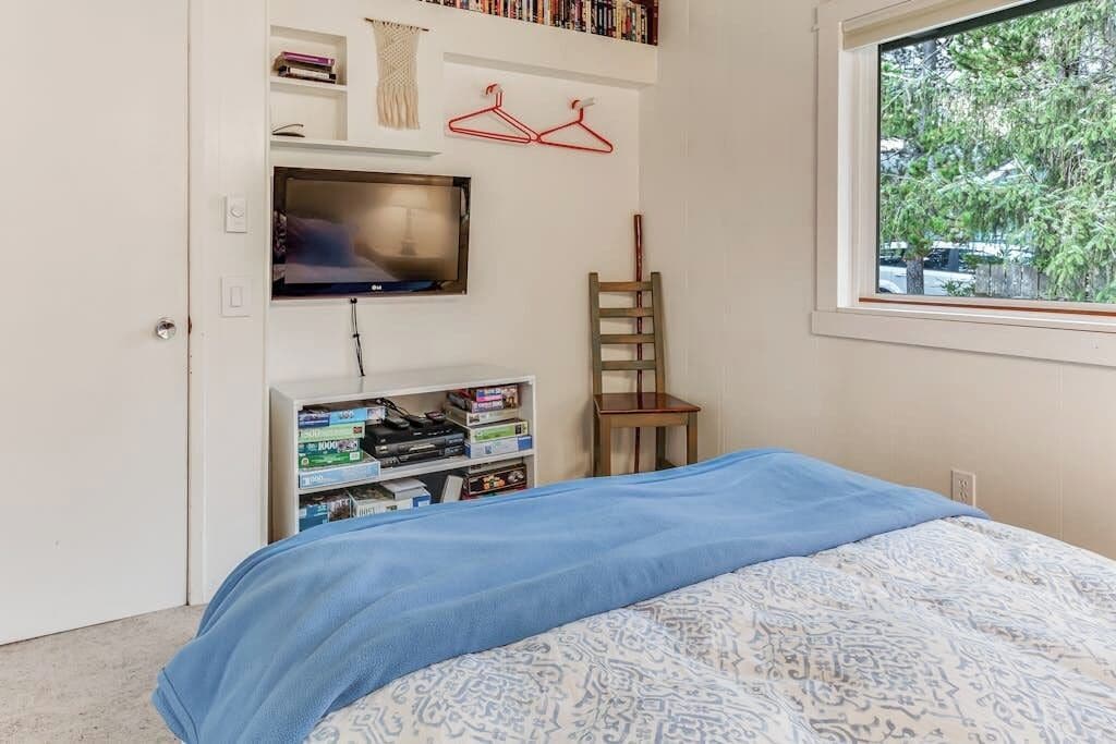 A bedroom with a bed, a television mounted on the wall, a bookshelf, and a window with a view of trees.
