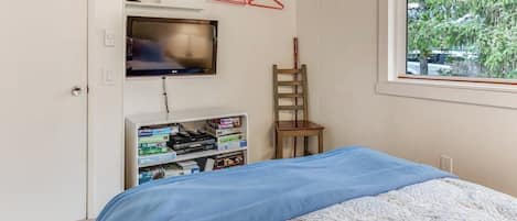 Un dormitorio con cama, televisor montado en la pared, estantería y ventana con vista a árboles.
