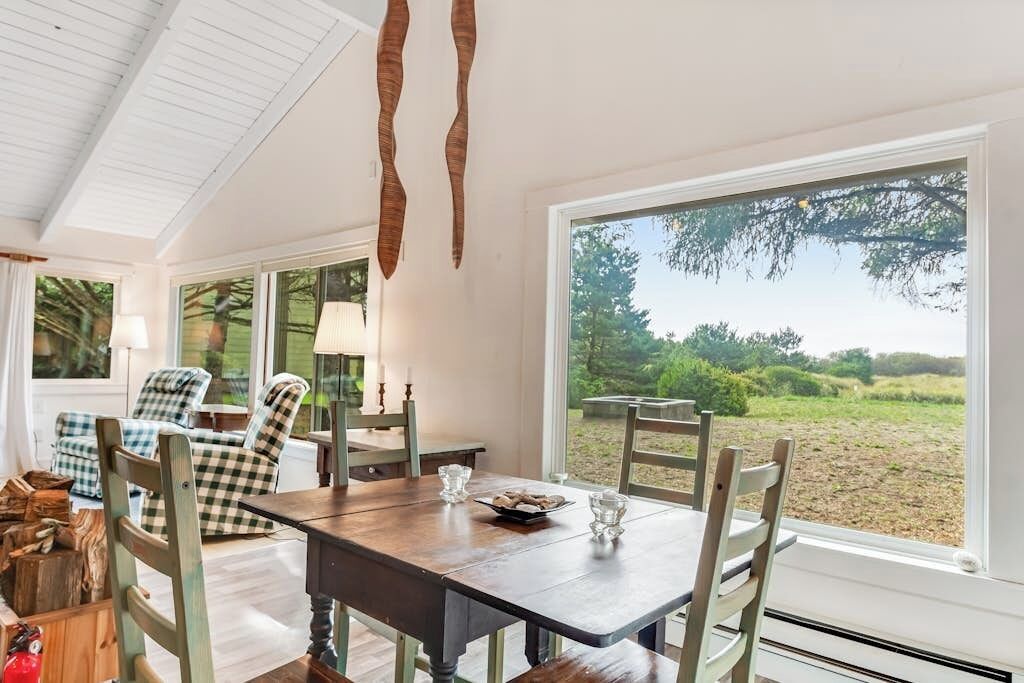 A dining area with a wooden table and chairs, large windows, and a view of the outdoors.