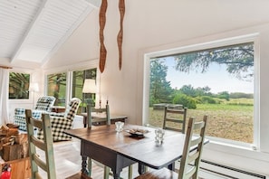 A dining area with a wooden table and chairs, large windows, and a view of the outdoors. - Adorable 2BR - WoodStove - Patio - Firepit - Washer Dryer (Copalis Beach)