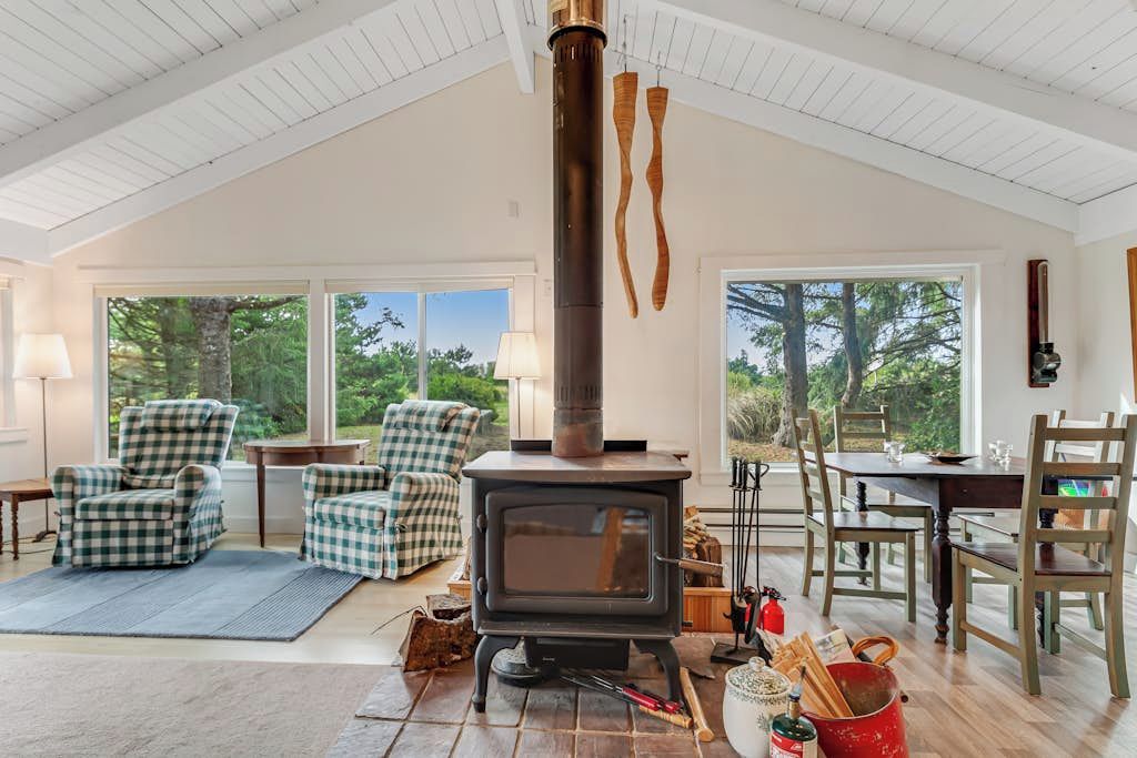A cozy room with a wood-burning stove, checkered chairs, a dining table, and large windows offering a view of trees.