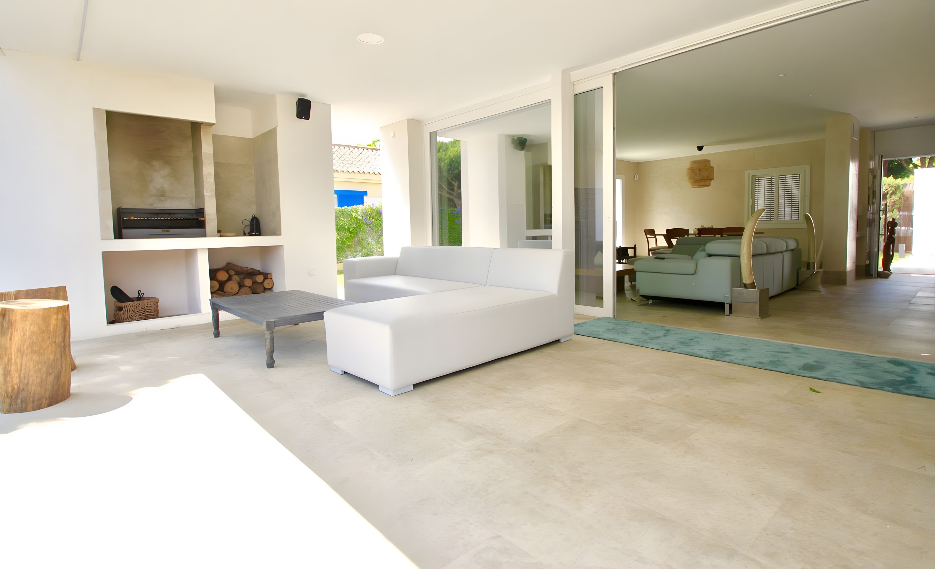 A modern living room with a white sofa, a coffee table, and a fireplace. There is a wooden table and a bench with a basket. The room has a large window and a view of the outdoors.