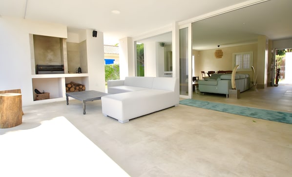 A modern living room with a white sofa, a coffee table, and a fireplace. There is a wooden table and a bench with a basket. The room has a large window and a view of the outdoors.