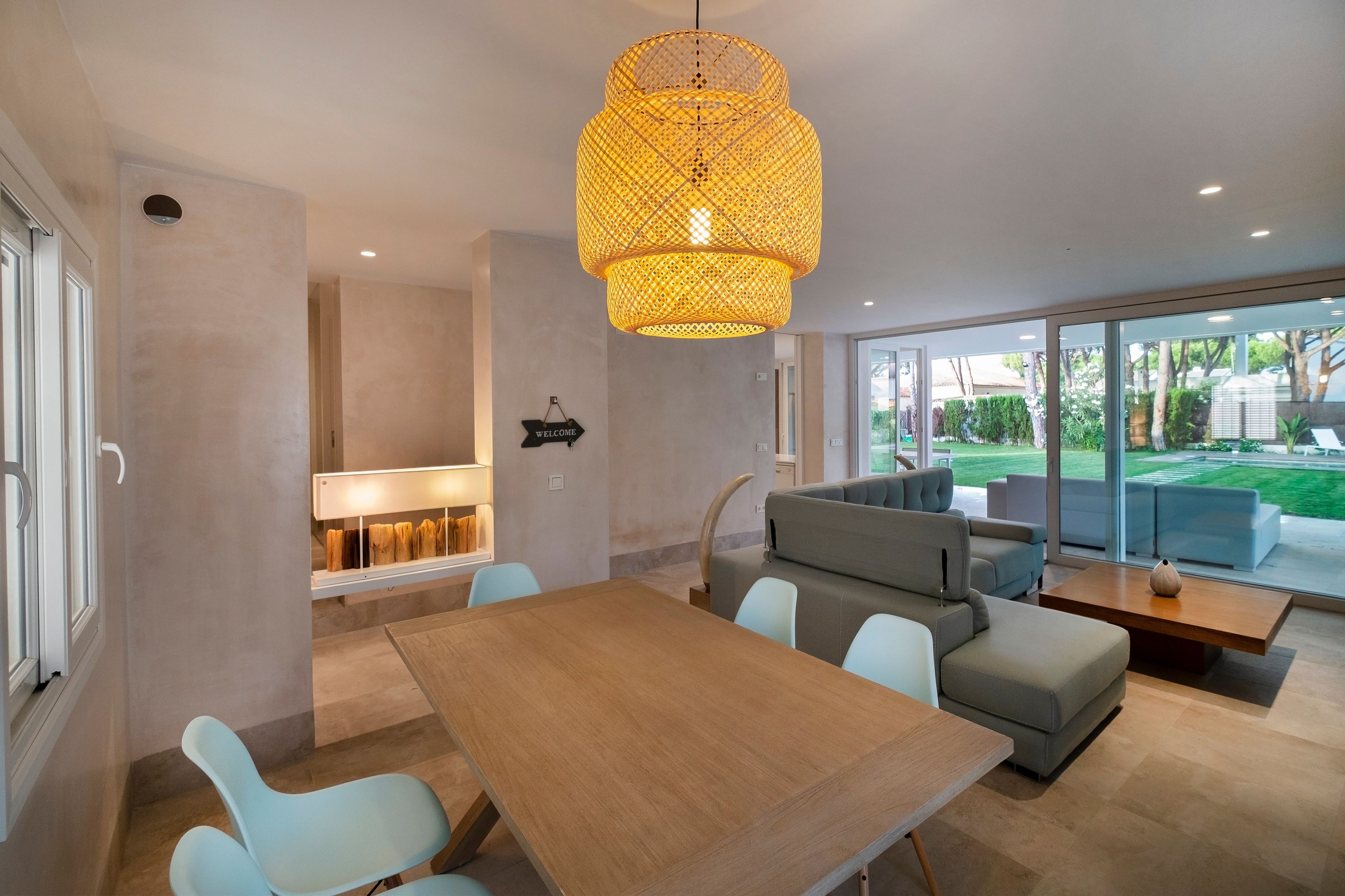 A modern living room with a wooden dining table, light blue chairs, a grey sofa, and a large woven light fixture.