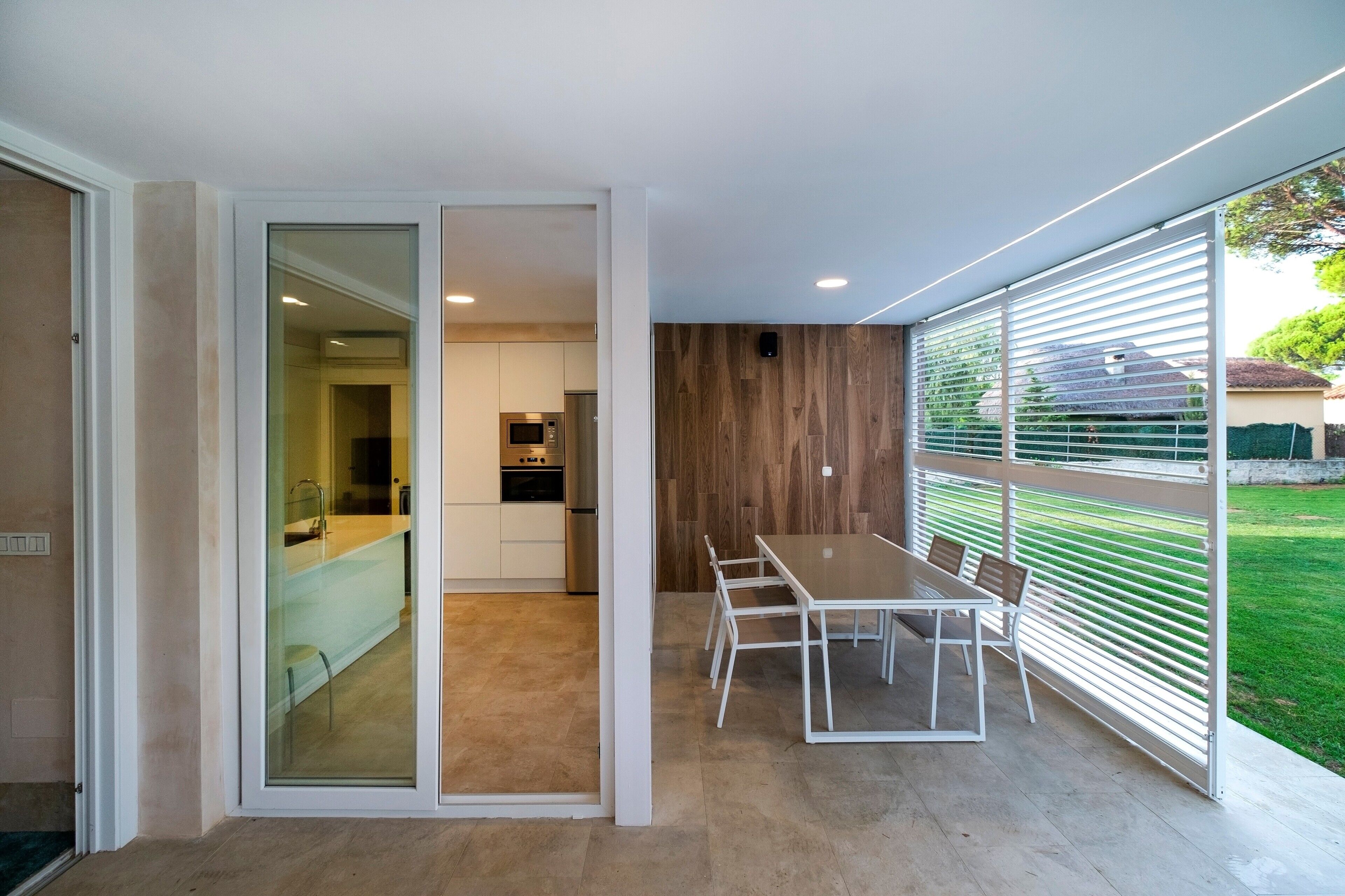 A modern kitchen with a built-in oven and microwave, a dining area with a table and chairs, and a view of a green garden through large windows.