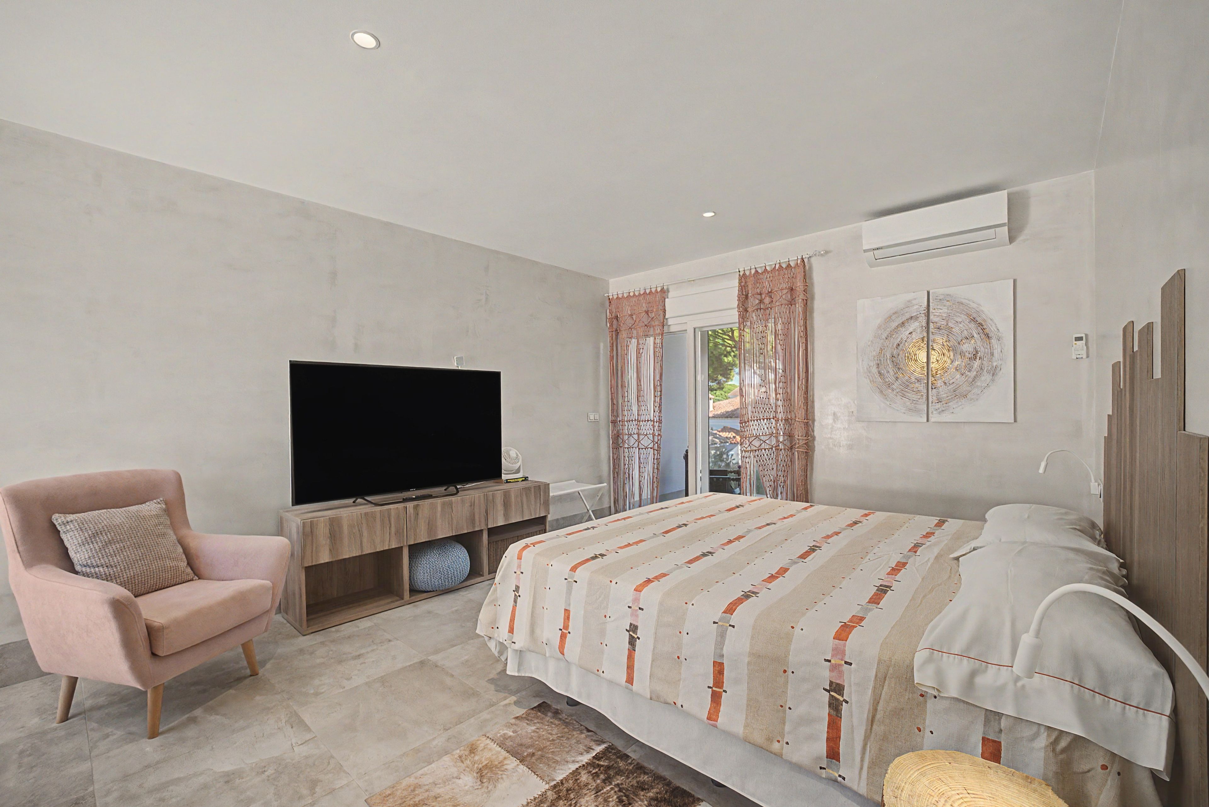 Een moderne slaapkamer met een flatscreen televisie, een bed met beddengoed met een patroon, een stoel en een groot raam met gordijnen.