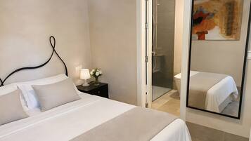 A hotel room with a bed, a nightstand, a vase with flowers, a glass shower enclosure, and a painting on the wall.
