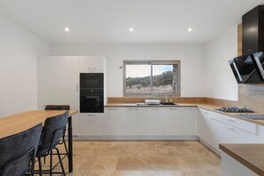 Una cocina moderna con armarios blancos, horno empotrado, ventana con vistas a árboles y un comedor con mesa y sillas de madera.