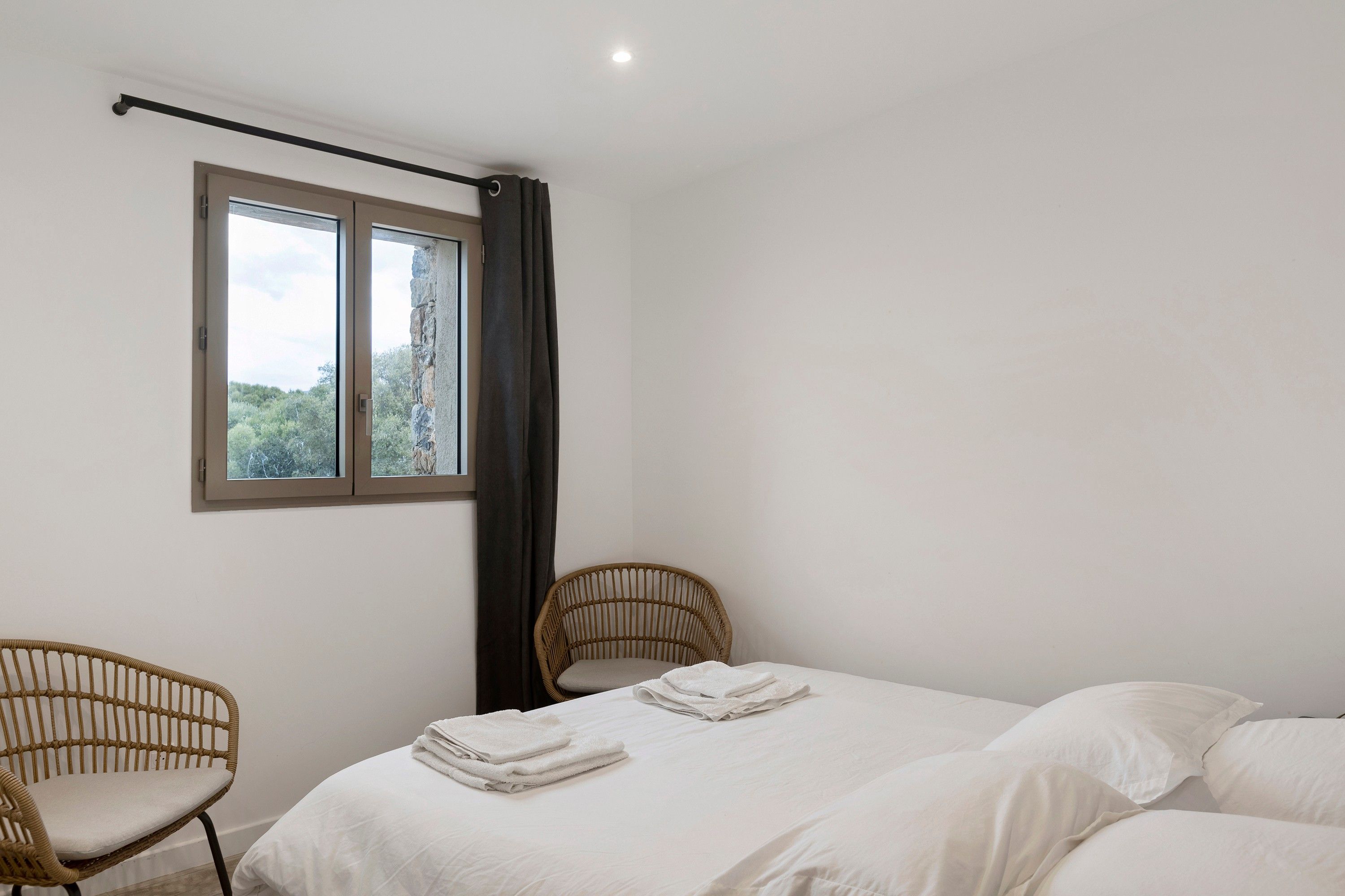 A bedroom with a bed, two wicker chairs, a window with curtains, and a view of greenery outside.