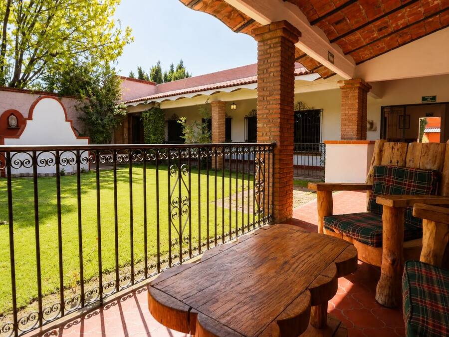 A covered outdoor seating area with a wooden table and chairs, a fenced yard, and a building with a red-tiled roof.