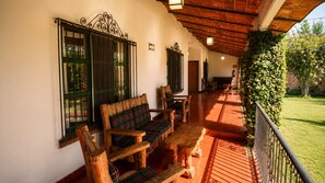 Un porche cubierto con mobiliario de madera, piso de baldosas y vista a un jardín.