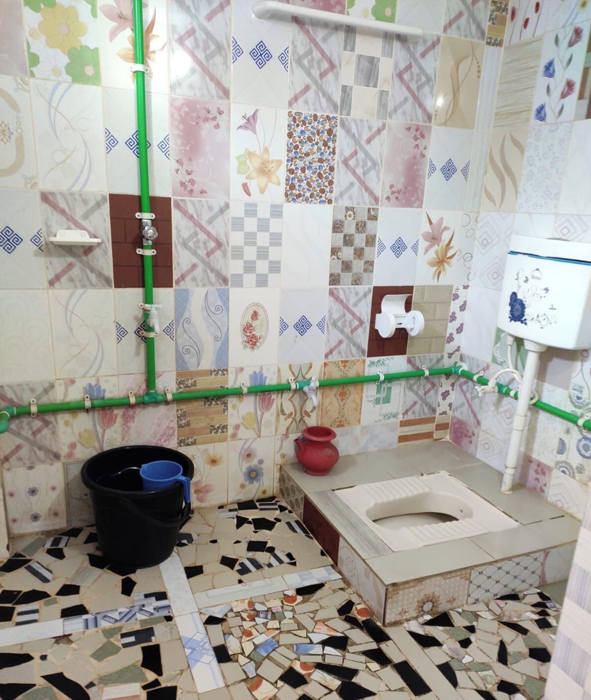 Een badkamer met een toilet, een emmer, een rode pot en een douchegedeelte met een groene leiding.